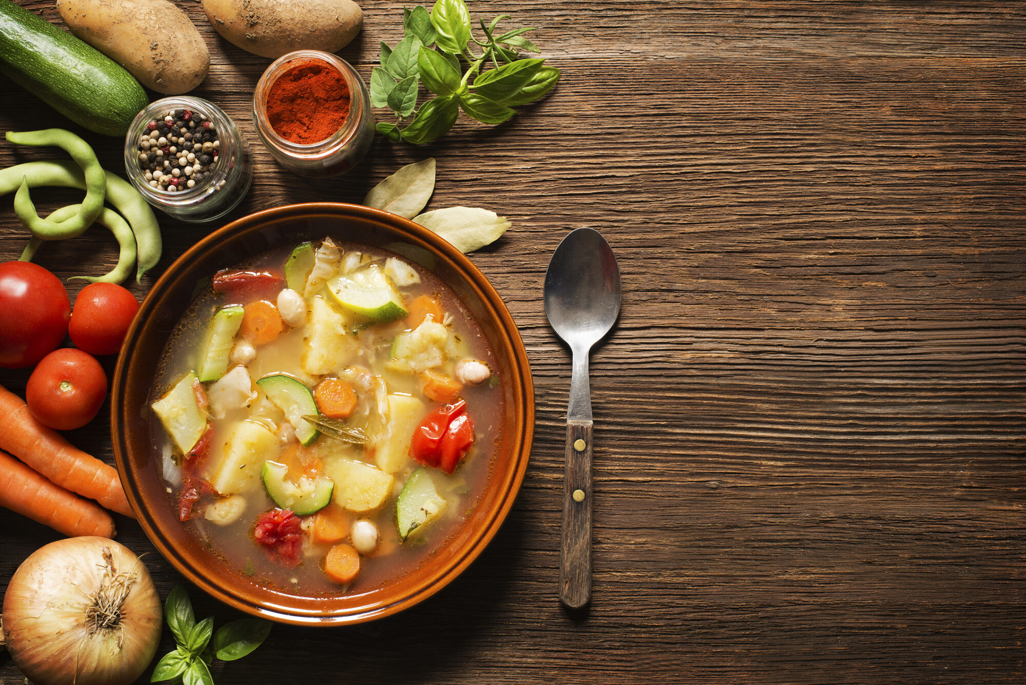 A bowl of fresh vegetable soup sits on a wooden table with the raw ingredients arrayed to one side.