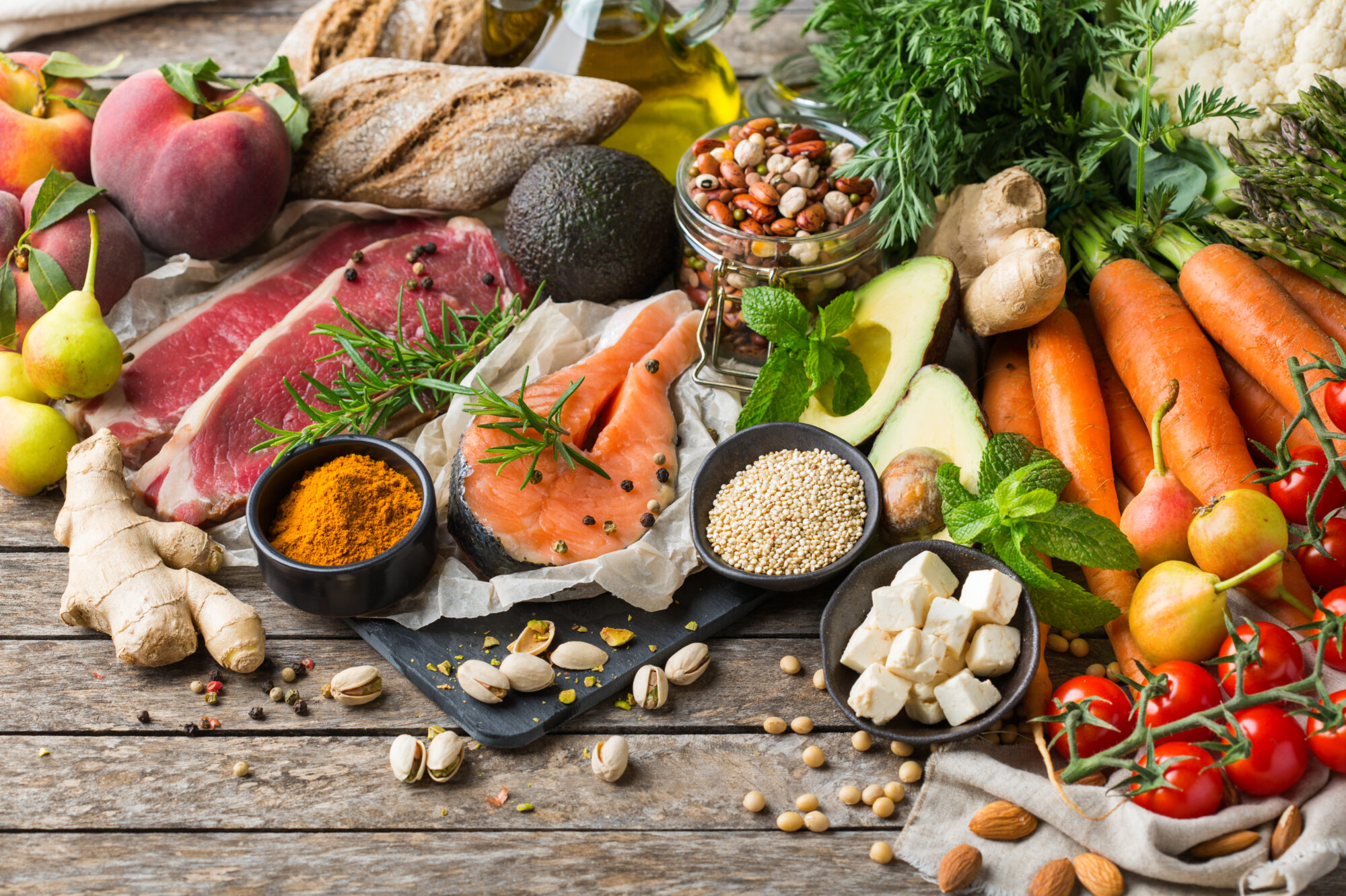 An assortment of healthy food ingredients for cooking on a wooden kitchen table, including vegetables, spices, nuts, legumes, and meats.