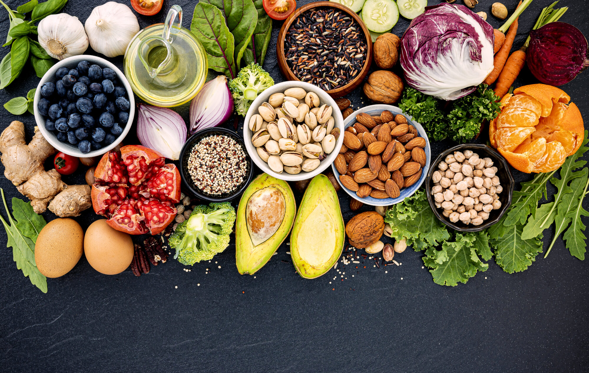 An assortment of vegetables, fruits, and legumes against a dark grey background. Bowls of pistachios, almonds, and dried chickpeas and wild rice sit next to an open avocado, raddichio, a jug of olive oil, fresh herbs and salad greens, and more.