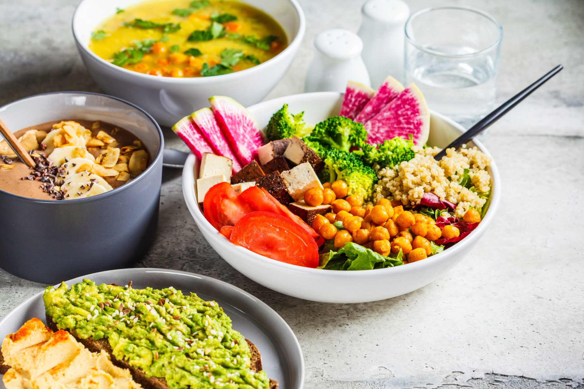 A close-up image of a colorful vegan lunch, with a rice bowl, bowl of soup, avocado toast, and chocolate smoothie bowl.