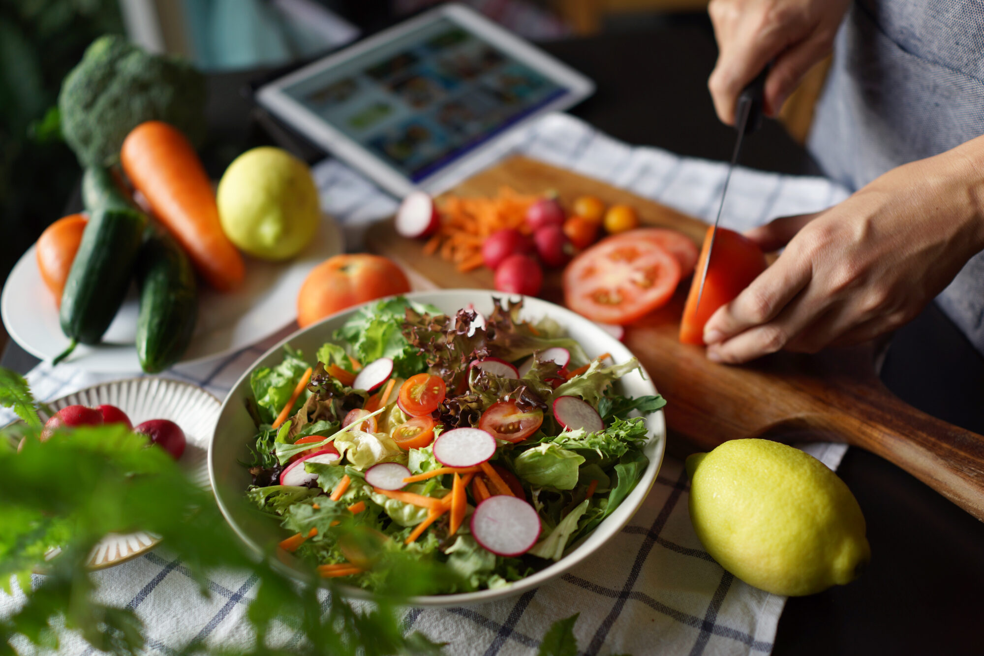 A close up image of a salad bowl with mixed greens, shredded carrot, radishes, and cherry tomatoes. In the background of the photo, someone is slicing a tomato while reading a recipe on a propped-up digital tablet.