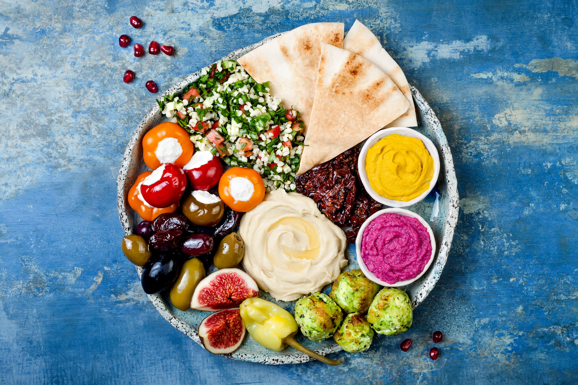 A Middle Eastern meze platter viewed from above, set on a blue table, with figs, dates, stuffed cherry tomatoes, hummus, pita bread, tabouleh, and more.