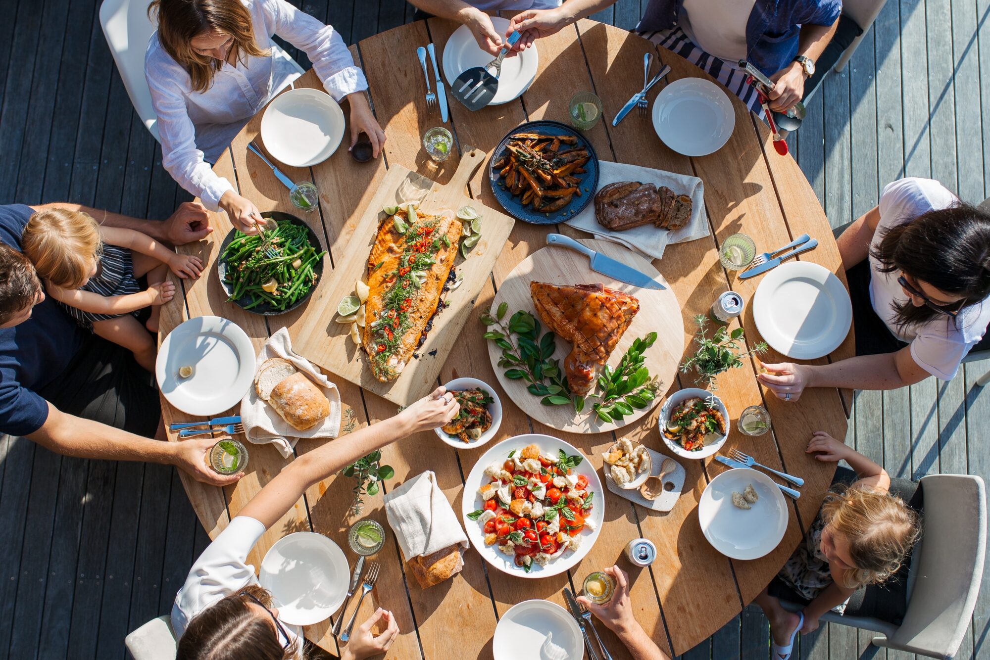 A view from above of nine people sharing a meal at a circular table. They are eating a colorful, fresh meal together with a caprese salad, a sie of glazed ham, fish, green beans, and more.